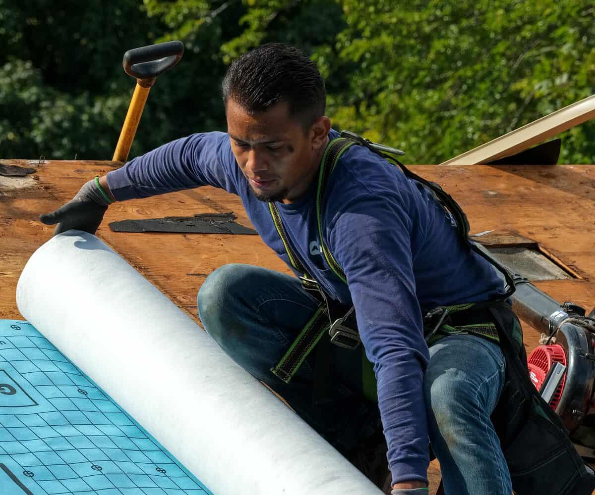Proliance roofer in a safety harness rolling out roof underlayment